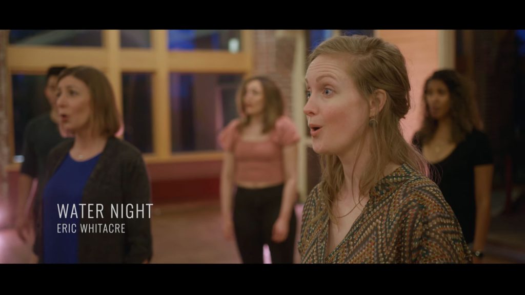 The words 'Water Night' on the screen with choral singers in the background against a windowed chapel